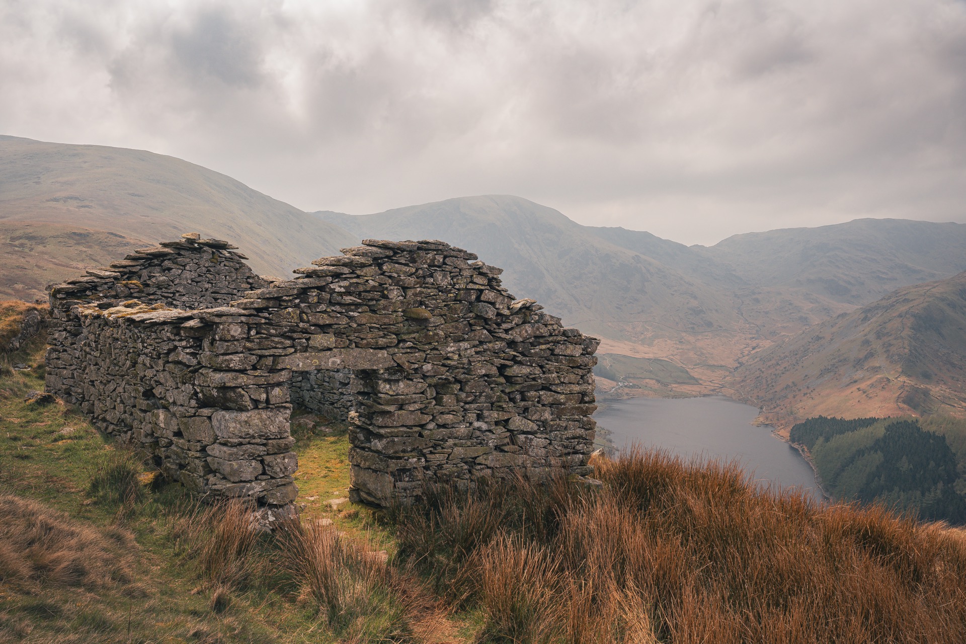 Dry stonewall building with a single door in the front. A missing roof but the front and back of the building clearly show the shape as it points up. Haweswater can be seen down below in the distance.